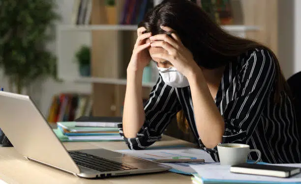 Sad teleworker woman wearing mask with problems due coronavirus sitting on a desk with laptop in the night at home office